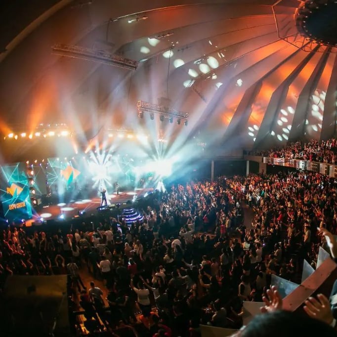 Público em casa lotada, com luz de palco e tela ao fundo